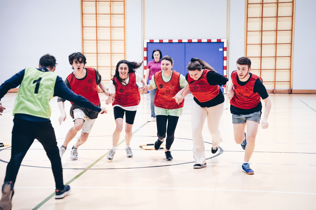Studenci biegną w strojach sportowych na hali sali gimnastycznej.